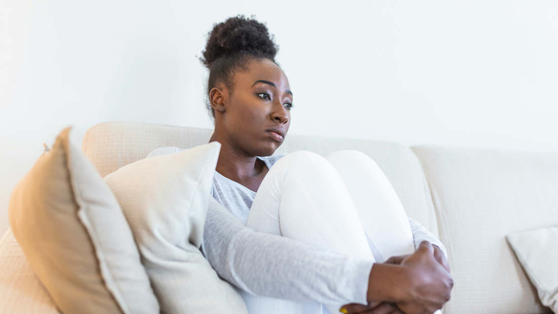 A young woman in a pair of white pajamas looks confused while sitting on a white couch holding her knees to her chest