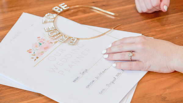 A hand with an engagement ring rests atop a wedding planning notebook. A headband that says “Bride To Be” lays nearby.