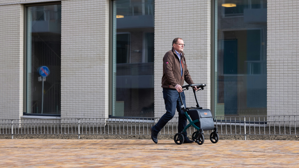 A man in a brown jacket wheels a scooter down a wide brick sidewalk past a white office building with three tall, narrow windows.