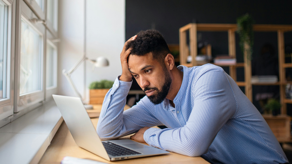 A man in a striped business shirt leans against a desk facing a sunlit window. He’s looking intently but defeatedly at a laptop.He’s leaned over the computer. One arm braces him against the desk. The other is propping up his head.