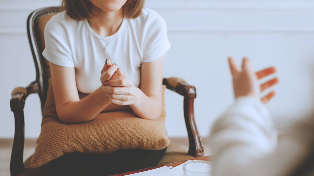 Một người ngồi trên ghế bành với một chiếc gối trên đùi, hai tay nắm chặt, trong khi một người khác ra hiệu về phía họ