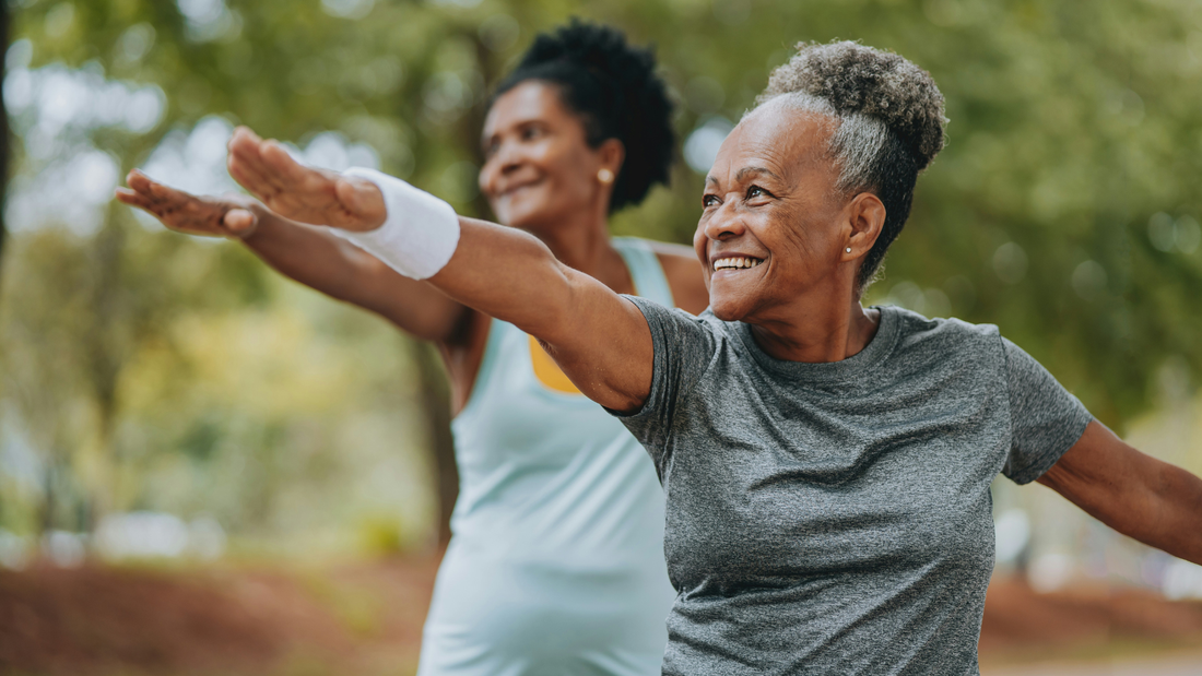 Two older women perform an aerobic workout in a park. Each has one arm outstretched before them and one arm stretching behind.