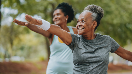 Two older women perform an aerobic workout in a park. Each has one arm outstretched before them and one arm stretching behind.