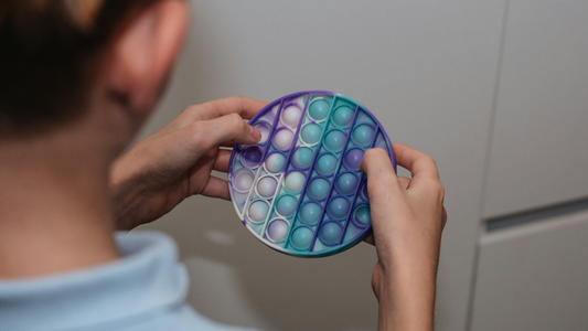 A child viewed from behind who is stimming using a popping toy.  The toy is blue, green, white and purple, and round in shape. 