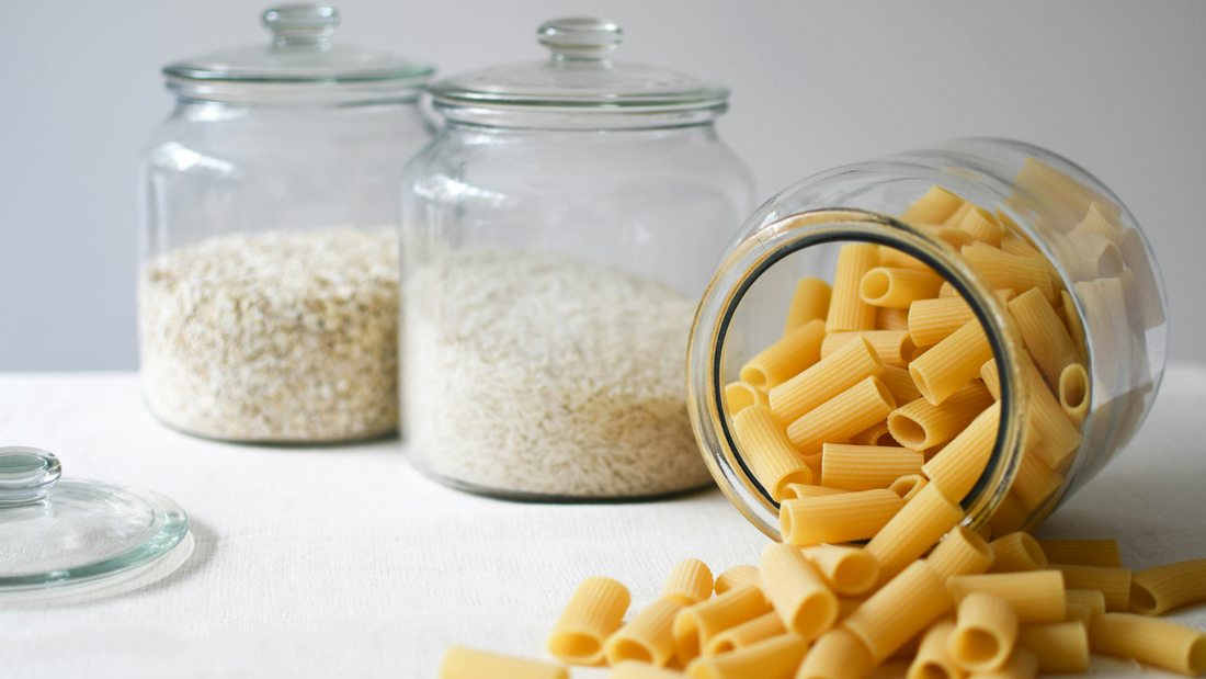 A row of three glass jars, each containing a grain. There is a jar of couscous, a jar of rice, and an overturned jar from which pasta is spilling out towards the camera.