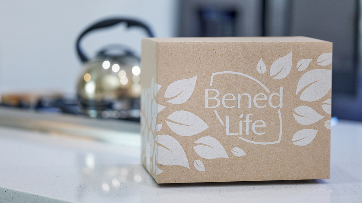 Một chiếc hộp Bened Life đặt trên mặt bàn màu trắng. Phía sau là một chiếc bếp có đặt ấm đun nước.