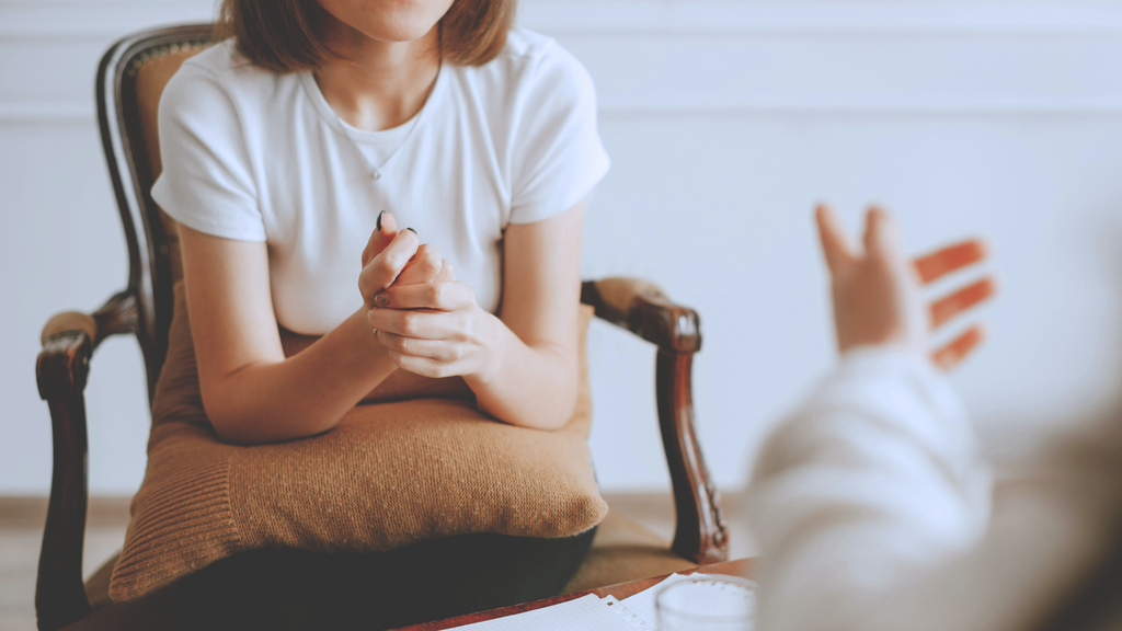 A person sits in an armchair with a pillow in her lap, hands clasped, as another person gestures toward them