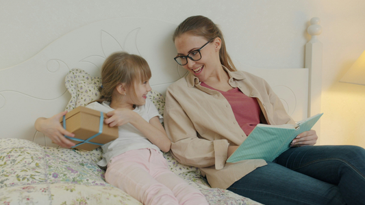 A mother and daughter recline in a bed together. The mother is reading a book and looking over at the smiling child, who is smiling back and holding a gift-wrapped box.