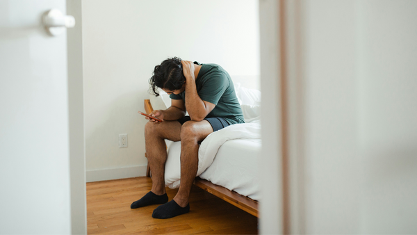 A person sits on the edge of a bed, cradling the back of their neck anxiously with their arm. They are focused on their phone, as if receiving bad news or doomscrolling.