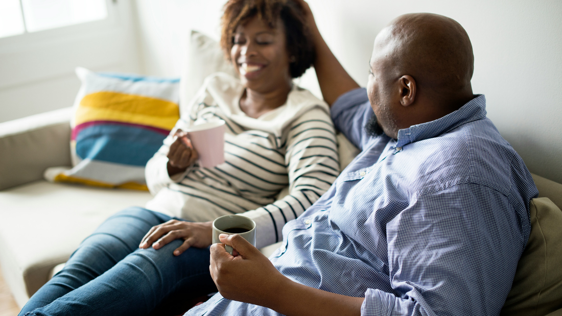 A couple sits on a sofa together, drinking coffee. The woman is grinning broadly, maybe even laughing. The man is turned toward her, his hand placed gently on the back of her head.