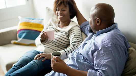 A couple sits on a sofa together, drinking coffee. The woman is grinning broadly, maybe even laughing. The man is turned toward her, his hand placed gently on the back of her head.