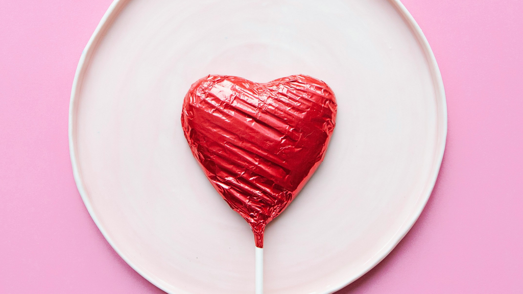 A heart shaped sucker in a red wrapper lying on a shallow pink dish on a darker pink surface