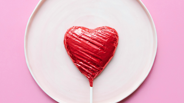 A heart shaped sucker in a red wrapper lying on a shallow pink dish on a darker pink surface