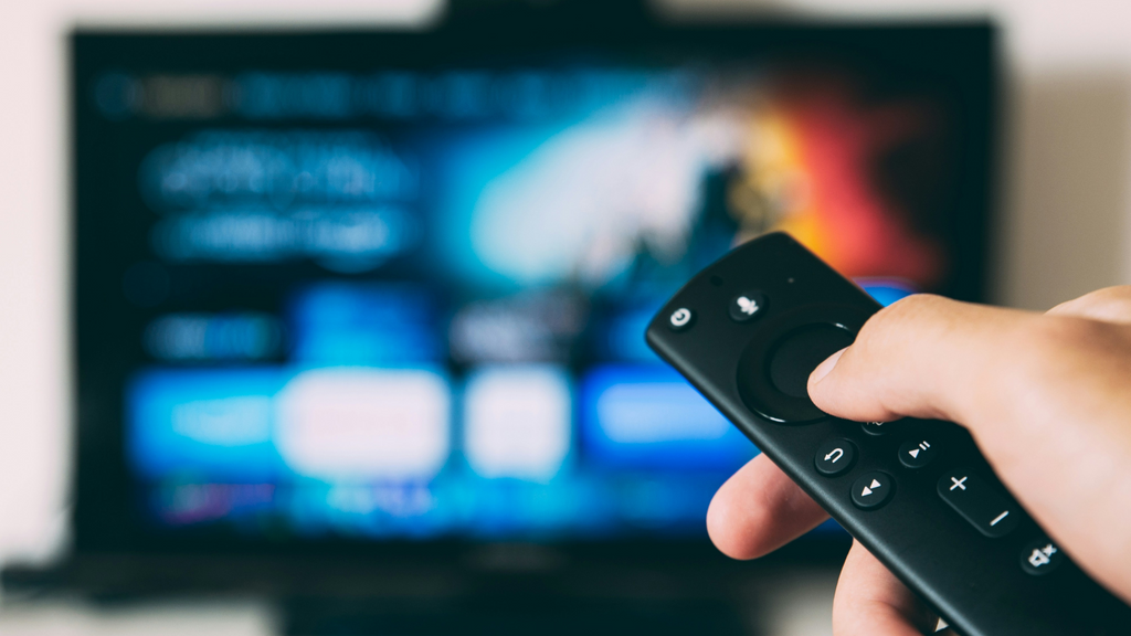 A finger on the button of a remote control with a TV tuned to a streaming service in the background.