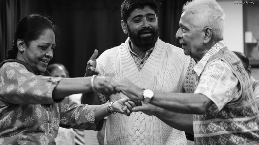 Black and white photo of Hrishikesh Pawar in a sweater vest. He is facing the camera, framed by an older couple who are holding hands, dancing in front of him.