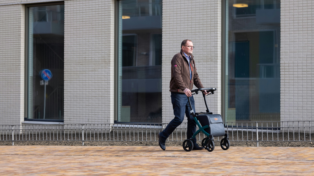 A man in a brown jacket wheels a scooter down a wide brick sidewalk past a white office building with three tall, narrow windows.