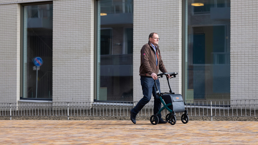 A man in a brown jacket wheels a scooter down a wide brick sidewalk past a white office building with three tall, narrow windows.