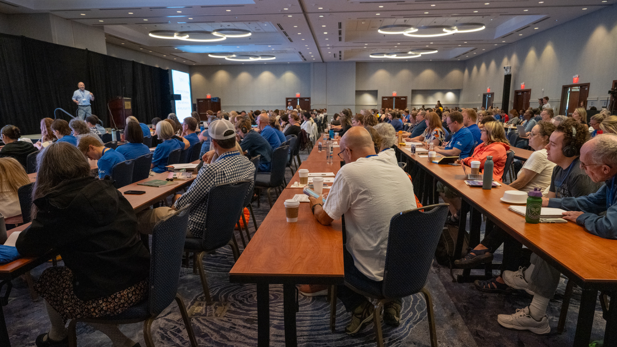 A lecture hall in a conference center. We are looking down long rows of tables where dozens of people are seated, paying attention to a speaker on stage to the left.
