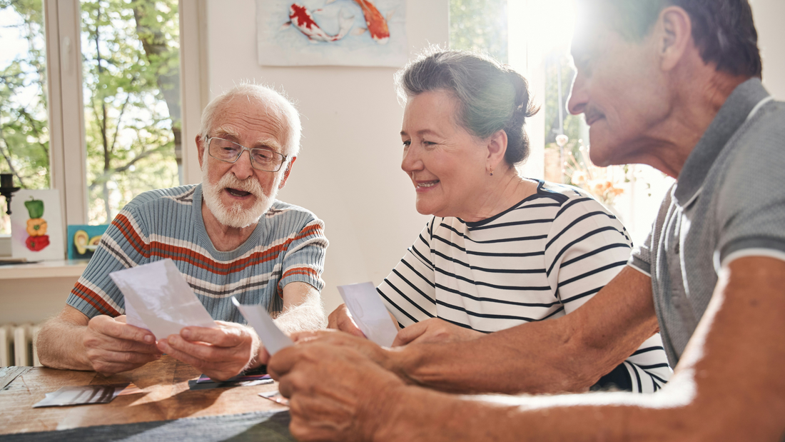 Three elderly people sit around a table sharing photographs with each other. A painting of koi hangs in the background between two windows.