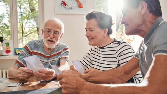 Three elderly people sit around a table sharing photographs with each other. A painting of koi hangs in the background between two windows.