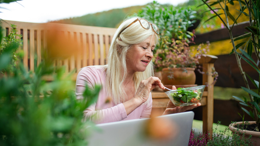 A middle aged woman with glasses on her head looks at a salad in a glass bowl while sitting outdoors amidst a variety of plants.