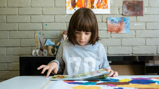 A child in a gray shirt looks down at a picture book. Colorful abstract fingerpainted pictures adorn the brick wall behind them and the table the book is lying on.