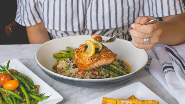 Someone seated at a table before a plate of grilled salmon, which rests on grilled asparagus and a bed of rice pilaf. A small forkful of salmon is in the person’s hand.