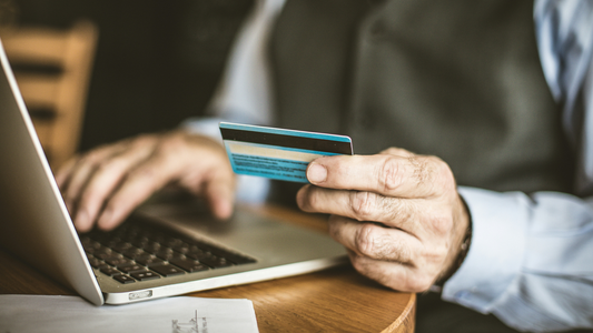 An older person sits at their laptop with a credit card in one hand. The other hand is resting on the keyboard. They are presumably about to make a purchase.