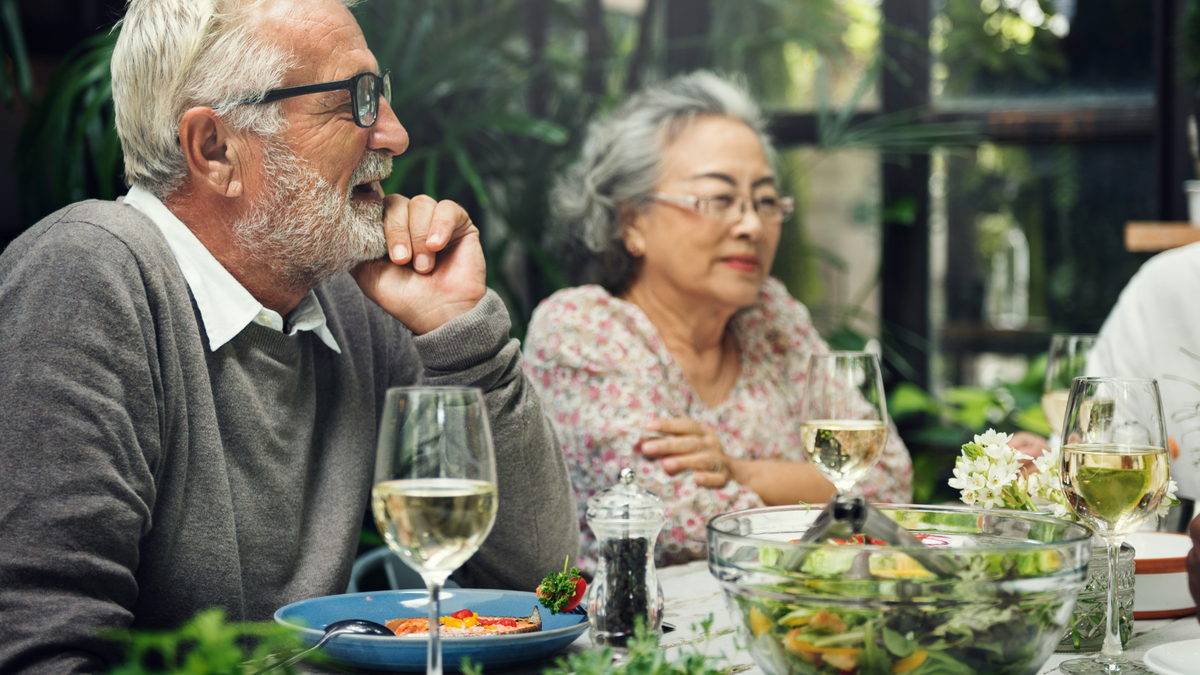 A senior couple at a nice dinner. There are dishes filled with fresh greens, glasses of wine, and lively conversation with others who are off-camera.