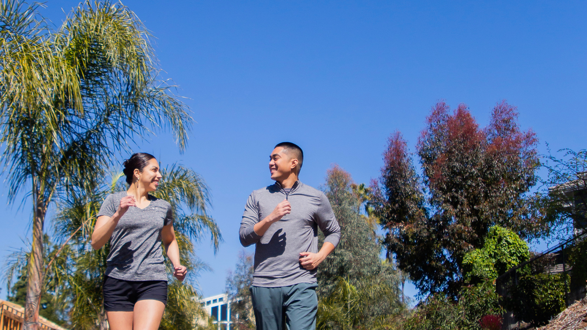 A man and a woman jog on a sunlit street. We are looking up at them; they are looking at each other and smiling. There is a palm tree in the background, and a clear blue sky beyond.