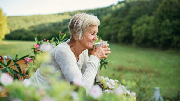 A senior woman sips a warm drink from a mug in a lush, green space. An arc of trees curves along the background and bright pink flowers frame her in the foreground.