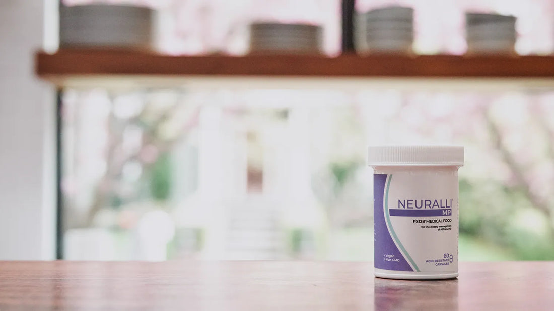 A bottle of Neuralli MP on a wood counter. Out of focus stacks of dishes stand behind it in front of a window, with trees outside.
