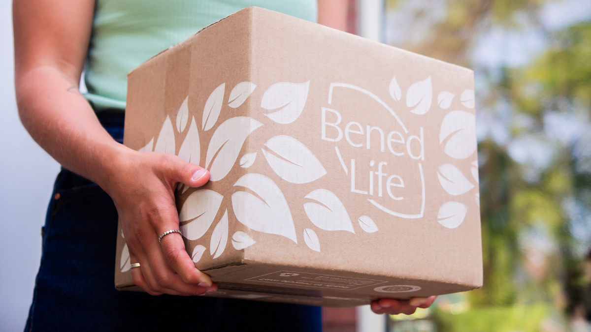 A woman in a short sleeved shirt and black pants holds a Bened Life box. Trees are out of focus in the background.