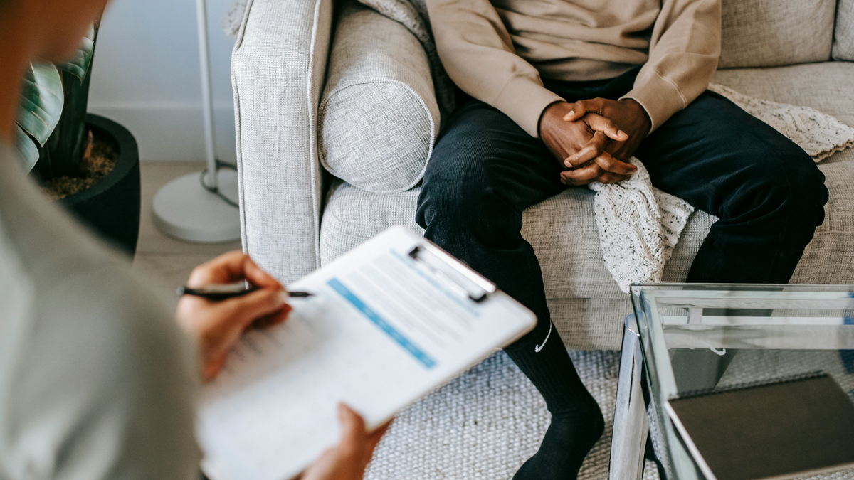 A person of color is seated on a couch opposite a mental health therapist who is holding a clipboard