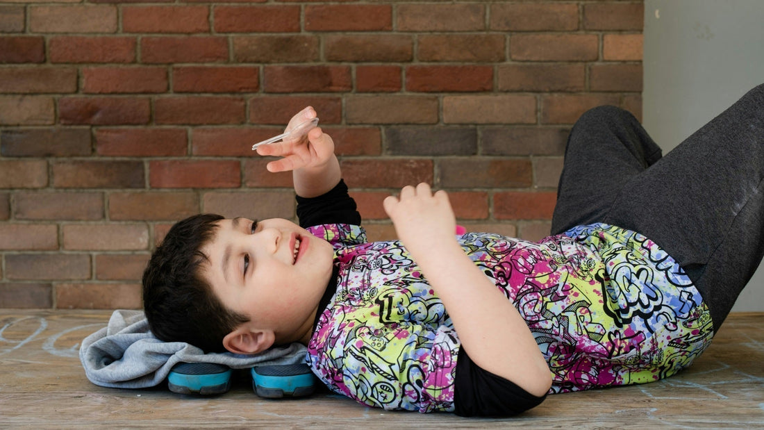 A child lays on a wooden floor on his back, playing. He is wearing a bright shirt with a graffiti-style art pattern