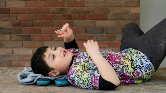 A child lays on a wooden floor on his back, playing. He is wearing a bright shirt with a graffiti-style art pattern
