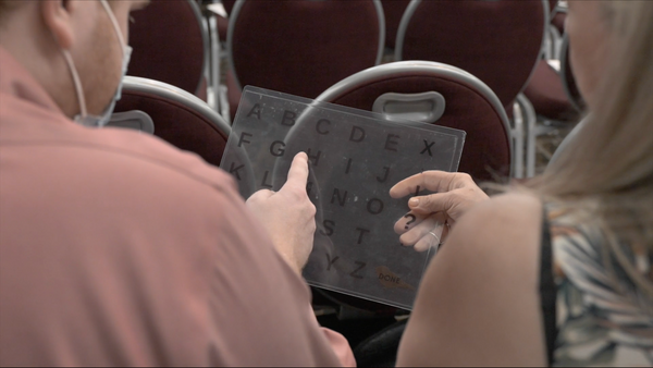 A person in a rose colored shirt points to a letter on a spelling-to-communicate board held by a woman sitting beside him.